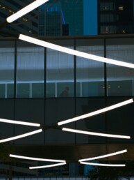 Crossed white light tubes in front of a glass office building at dusk
