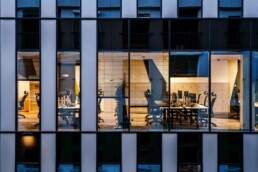 Lit open-plan office with desks and chairs seen through a building's windows at dusk