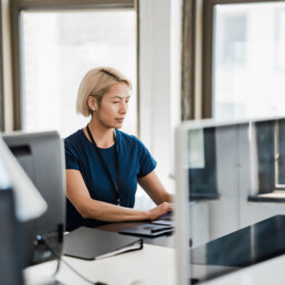 Woman in a blue shirt typing on a laptop at a desk beside a large window