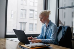 Woman in a blue shirt typing on a laptop at a desk beside a large window